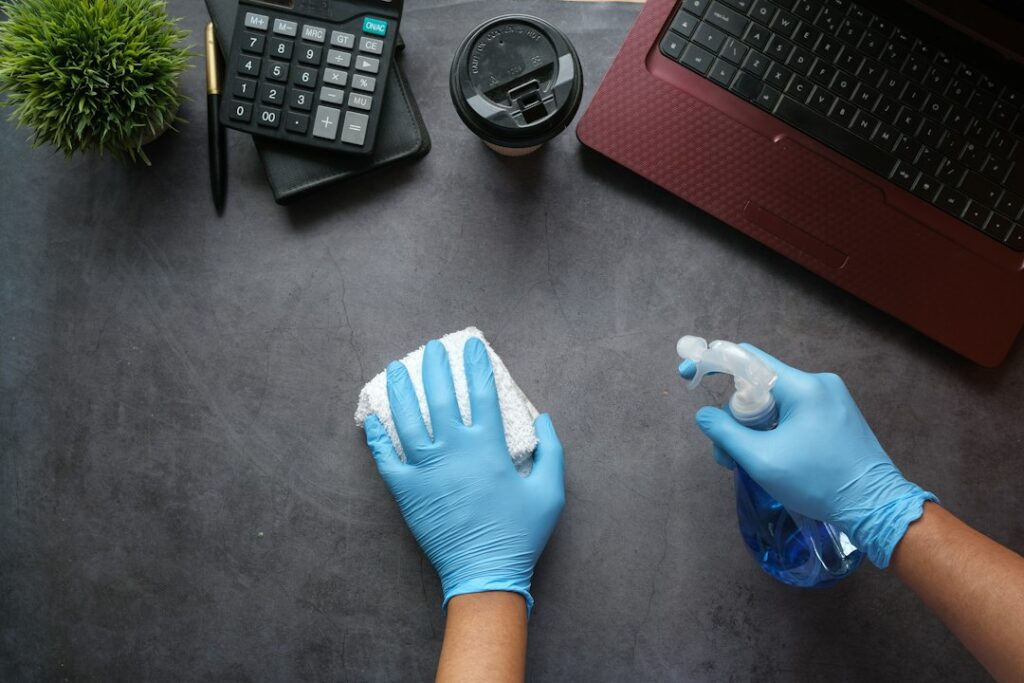 Mani con guanti blu puliscono una scrivania con spray e panno, pratica collegata all'individuazione del coronavirus sulle superfici