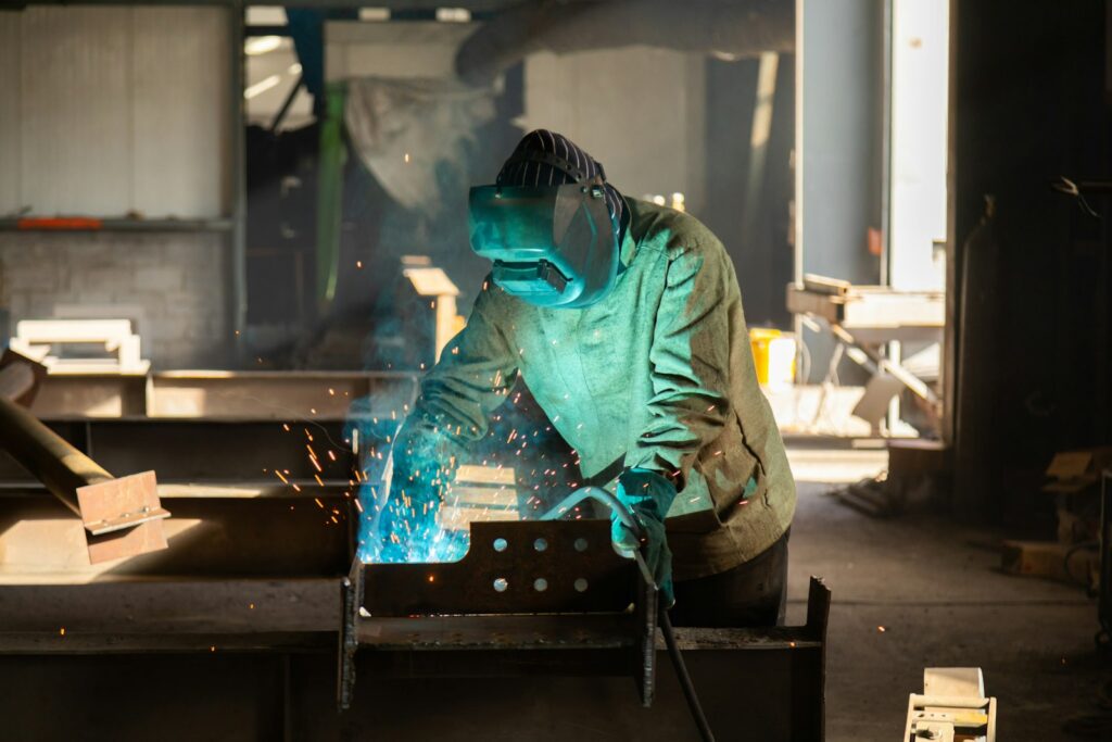 Operaio saldatore con casco protettivo e torcia che salda profili metallici in officina, investimenti in sicurezza nelle PMI