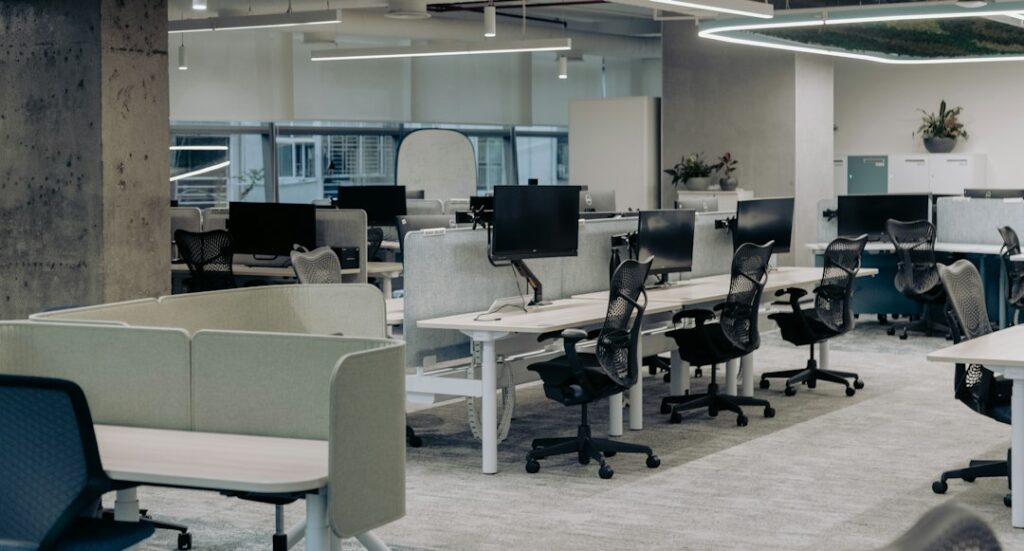 Ufficio open space con scrivanie, computer e sedie ergonomiche separate da divisori bassi, ambiente da analizzare nella valutazione dello stress da lavoro correlato